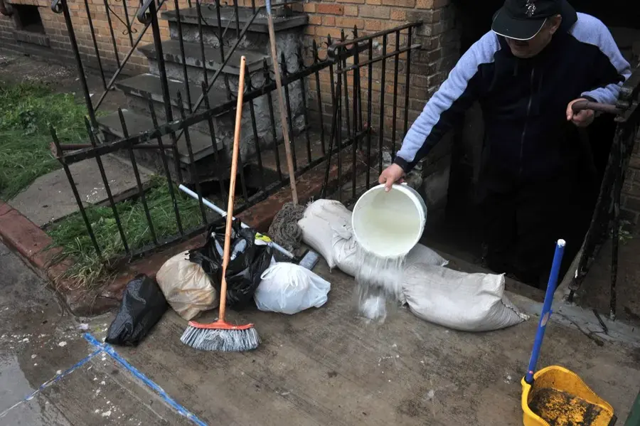 Resident bailing storm water beside sandbags at a brick rowhouse basement stair